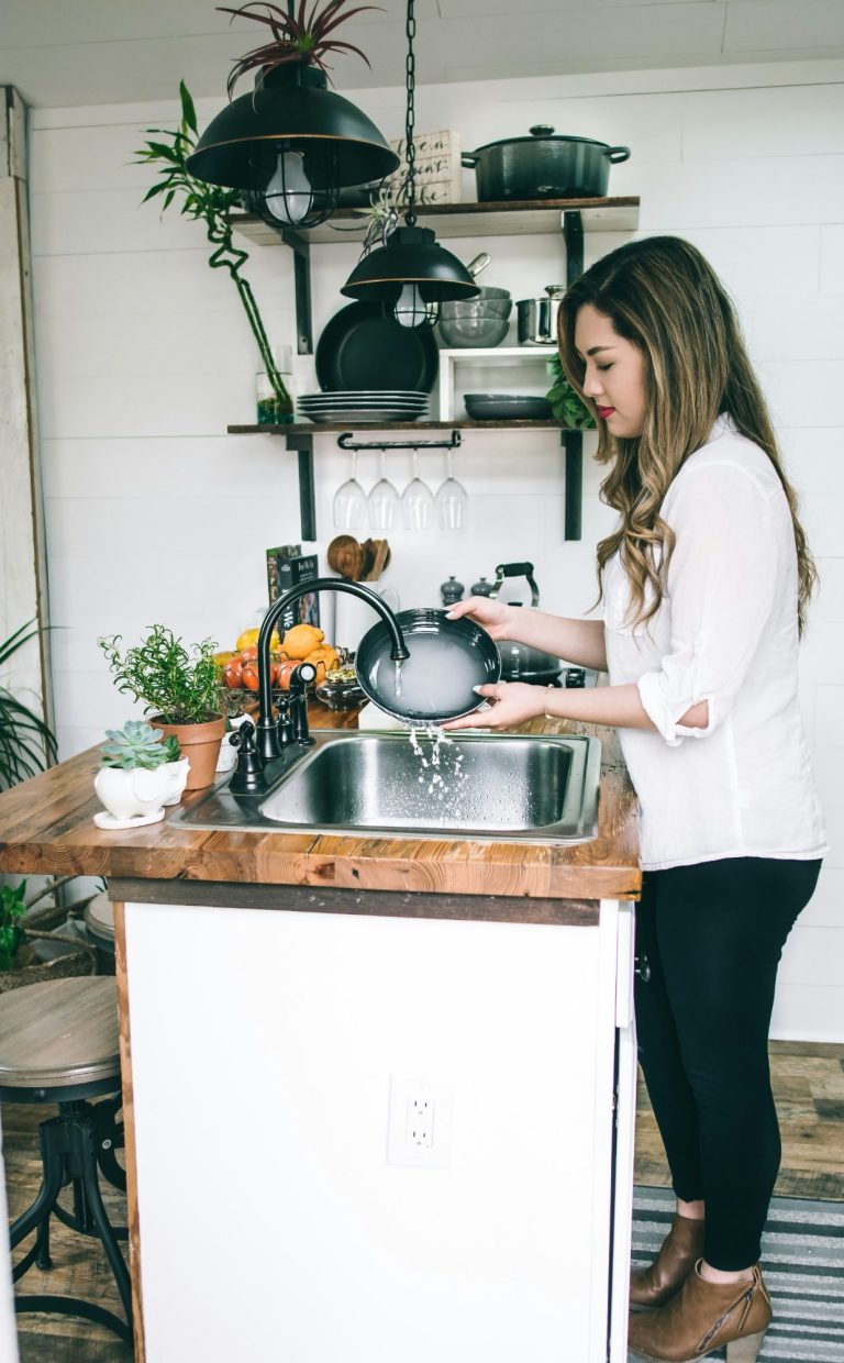 Person washing dishes in a modern student kitchen with houseplants and shelves, representing easy cleaning tips for student accommodation.